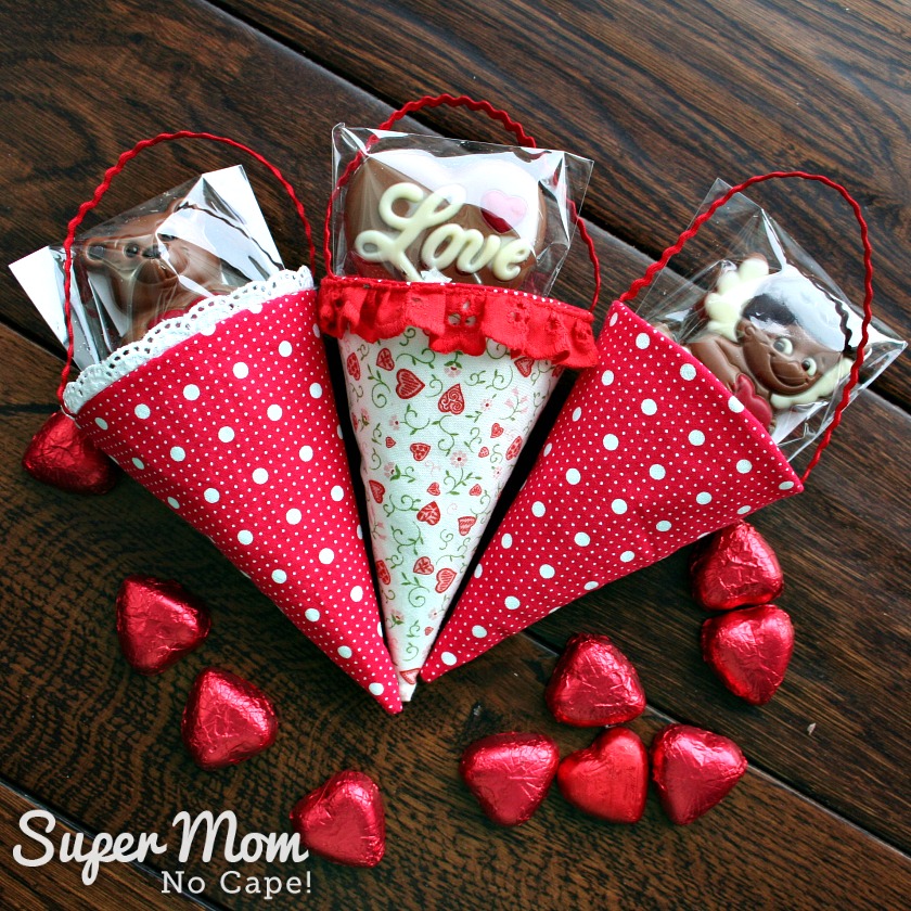 Photo of three fabric cones filled with Cadbury hearts and chocolate suckers laying on a dark wood table with red heart shaped foil wrapped candy scattered around.