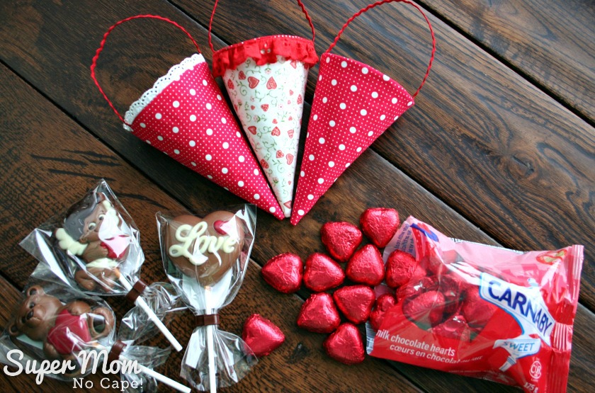 Photo of three empty fabric cones on a dark wood table with Cadbury heart and chocolate suckers and red heart shaped foil wrapped candy spilling out of a bag.