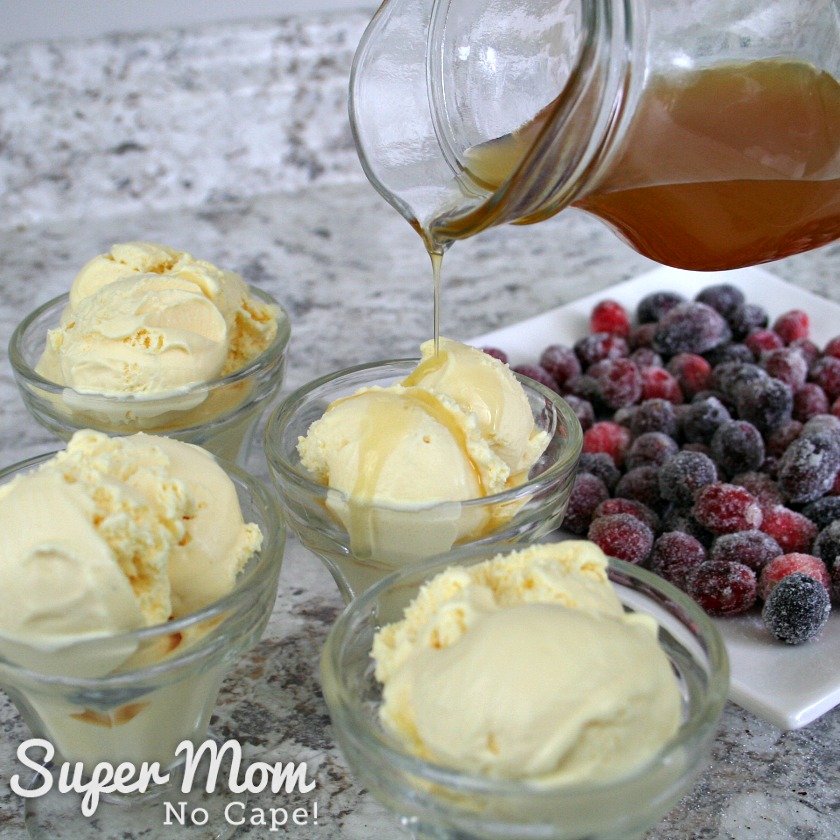 Ice cream in bowls with orange sauce being drizzled on top with plate of sugared cranberries in the background