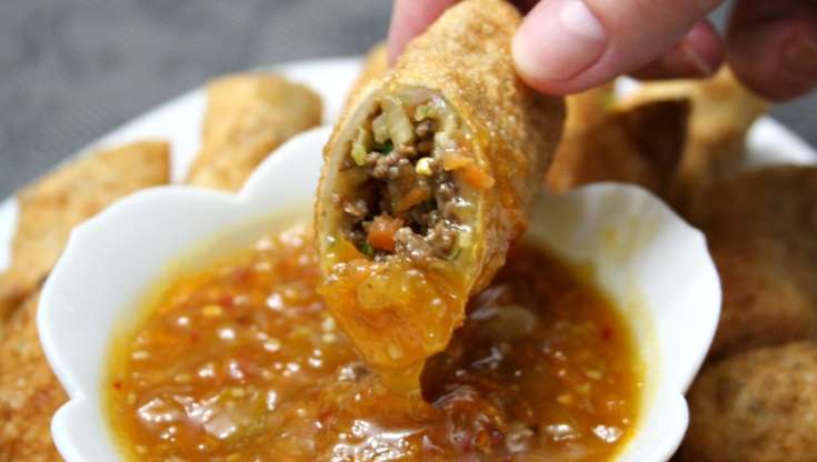 White bowl with orange sauce and egg roll being dipped into it