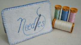 White felt needlebook with Needles embroidered on it standing beside 4 spools of thread