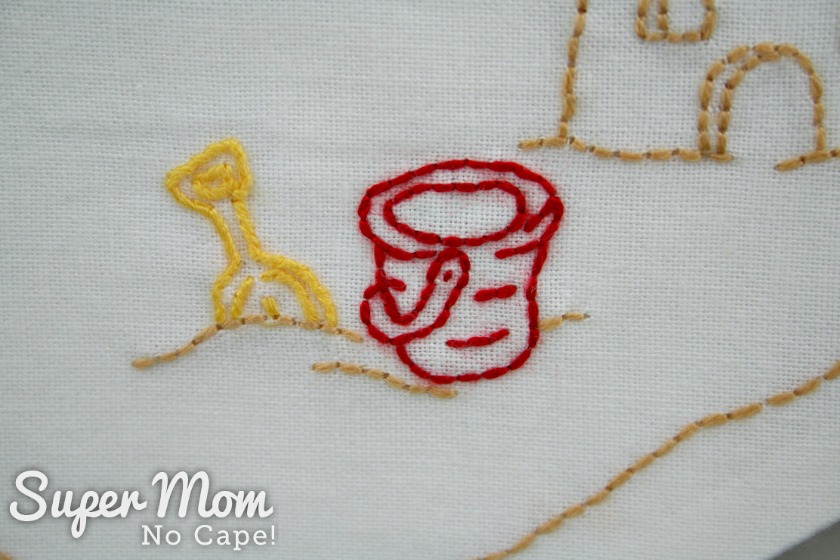 Embroidered red bucket and yellow shovel sitting on sand beside sand castle