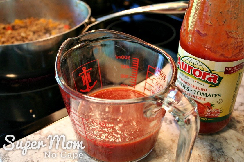 Measuring cup with one cup of crushed tomatoes beside a jar of crushed tomatoes with frying pan in the background