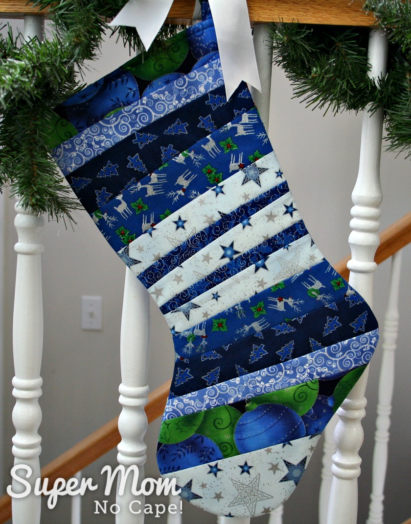 Blue quilt as you go stocking with toe facing left tied with ribbon to a banister wrapped in pine garland.