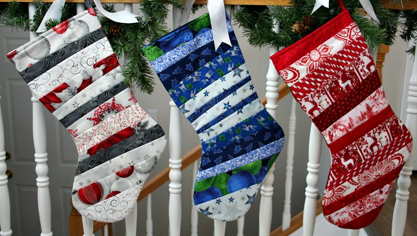 Three quilt as you go stockings hung on a handrail wrapped with pine branches
