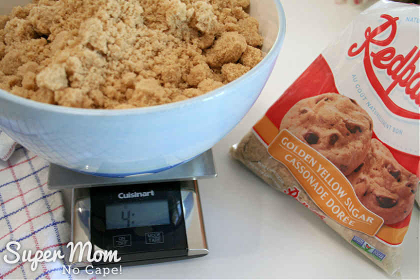 Blue mixing bowl full of brown sugar on a kitchen scale beside a bag of brown sugar.