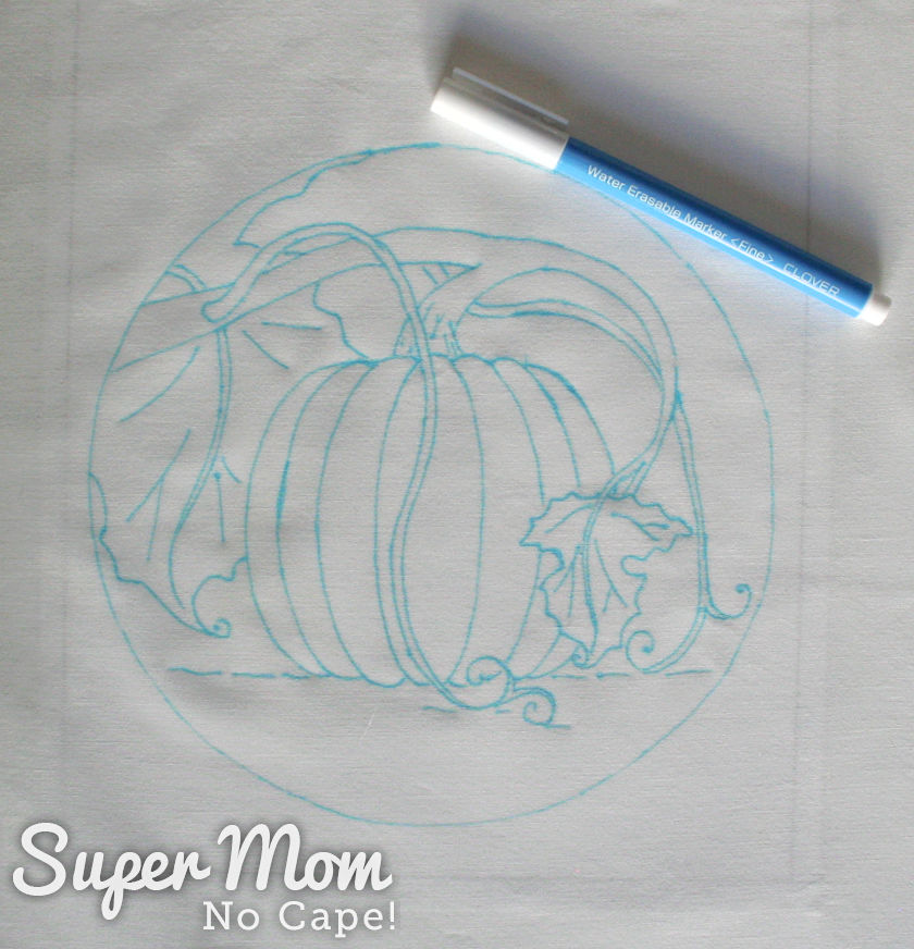 Pumpkin embroidery traced onto light colored fabric and a blue water soluble maker beside it.