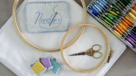 A large and small embroidery hoop, needle book, scissors and floss bobbins on white fabric beside a floss storage container.