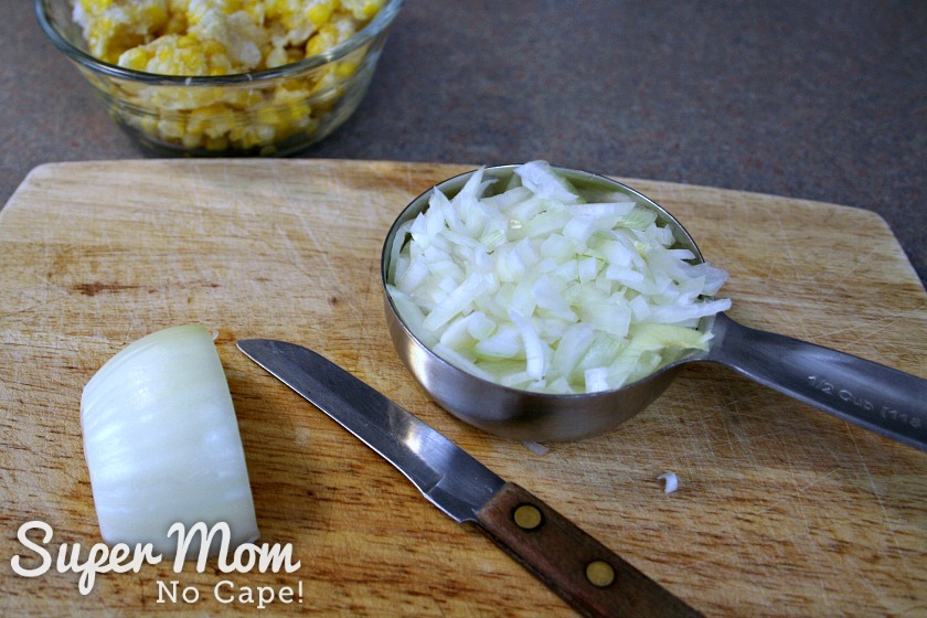 A picture of diced onions in a 1.5 cup measuring cup., with an additional half onion, unchopped to the left, on a cutting board. A bowl of frozen corn kernels is in the background.