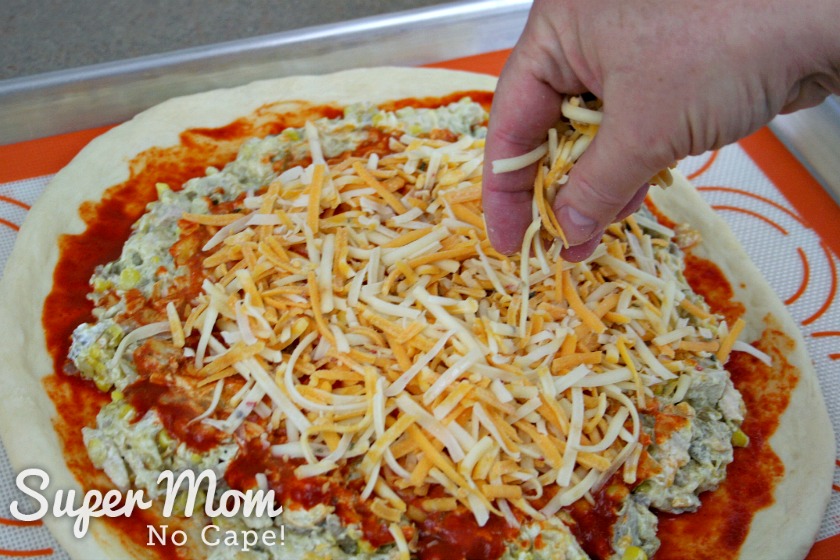 A picture of a person's hand spreading the shredded cheese over the filling that has been covered with enchilada sauce.
