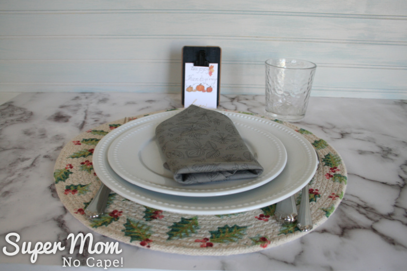 Thanksgiving place card sitting behind a place setting with a gray napkin. The place card has pumpkins.